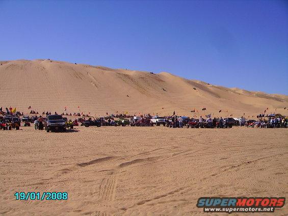 Desert dunes with many people and off-road vehicles gathered on a sunny day