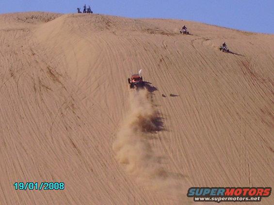 Orange dune buggy descending a sand dune, kicking up dust. Three ATVs are visible on the dune crest