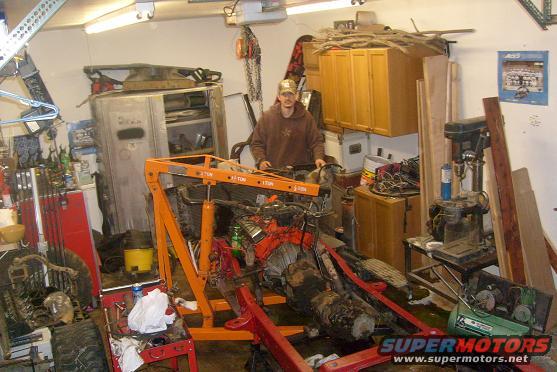 Man in garage with engine on an orange engine hoist, surrounded by tools and storage