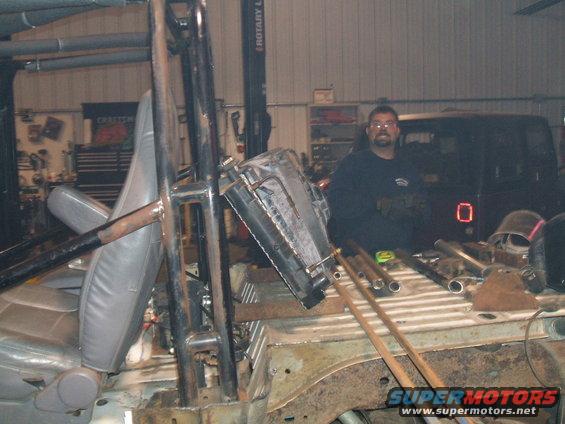 man standing in a garage next to a disassembled vehicle, with a radiator and metal tubes on the floor