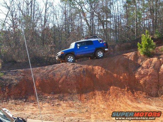 Blue Toyota FJ Cruiser climbing a steep dirt hill with trees in background. Roof rack visible