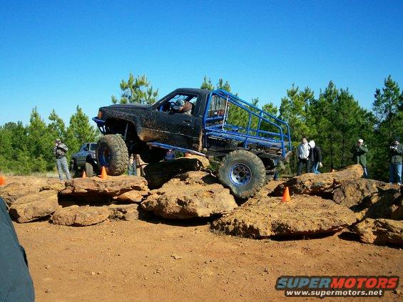 Modified off-road truck with blue roll cage climbing over rocks. Another SUV is visible behind it
