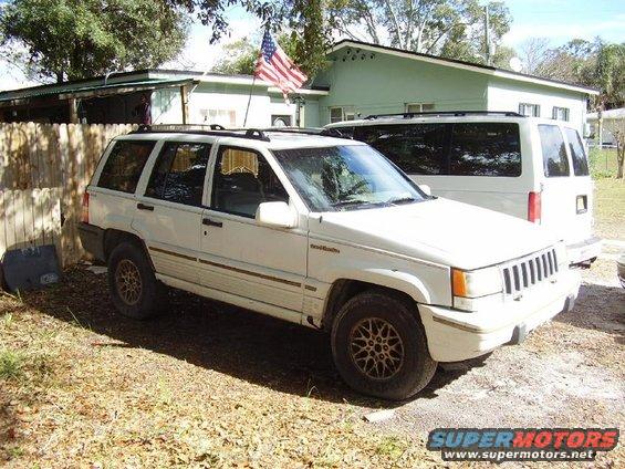 White Jeep Grand Cherokee parked in a yard next to a white van. American flag flying on a pole