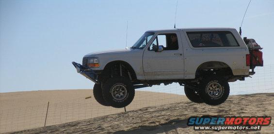 White Ford Bronco airborne over sand dune, modified with off-road suspension and bumper