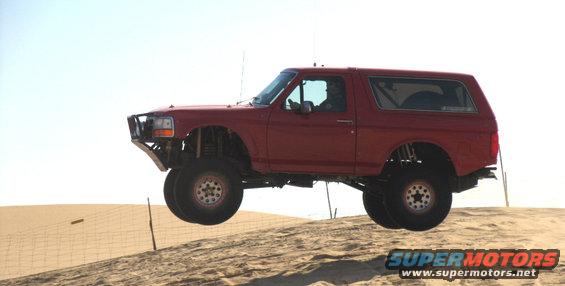 Red Ford Bronco mid-air jump over sand dune, aftermarket bumper, lifted suspension