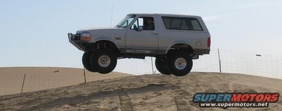 Silver Ford Bronco mid-air over a sand dune, lifted suspension, off-road tires