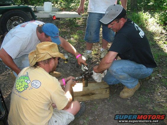 Three men gathered around a mechanical component on a wooden block outdoors