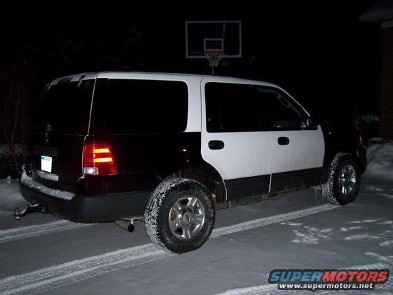 SUV with black rear and white front parked in snow at night. Basketball hoop in background