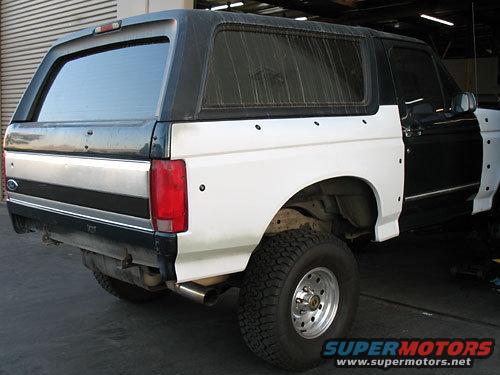Rear quarter view of a black and white Ford Bronco with primer-white fenders, in a garage