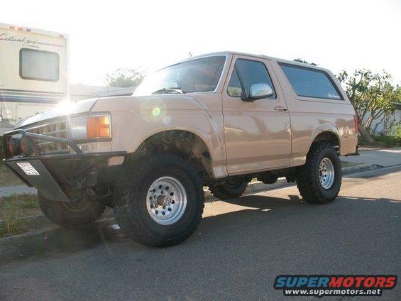 Tan 1987 Ford Bronco with aftermarket bumper and large tires parked on a street