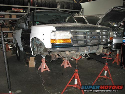 Front view of a dark green Ford Bronco, undergoing repairs, with white fender flares and disassembled front end