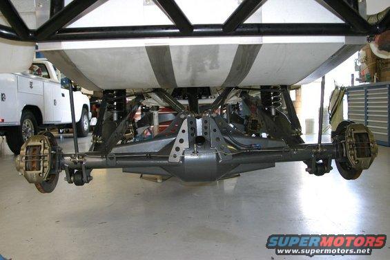 Underside view of a vehicle's rear axle and suspension assembly, with a white service truck in the background