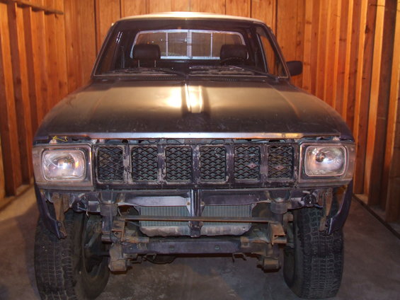 Front view of a dark-colored Toyota pickup truck, possibly from the 1980s, with a damaged grille and bumper