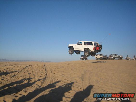 White 1995 Ford Bronco catching air over sand dunes with other vehicles in background