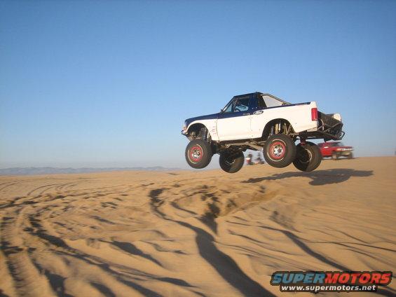 lifted pickup truck mid-air over sand dunes, another car in background
