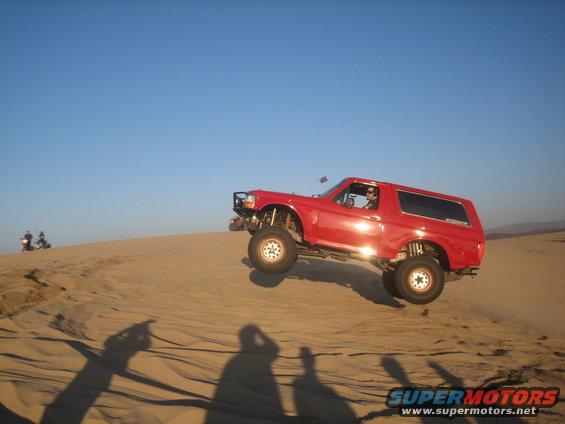 Red Ford Bronco mid-air over a sand dune, lifted suspension, aftermarket bumper