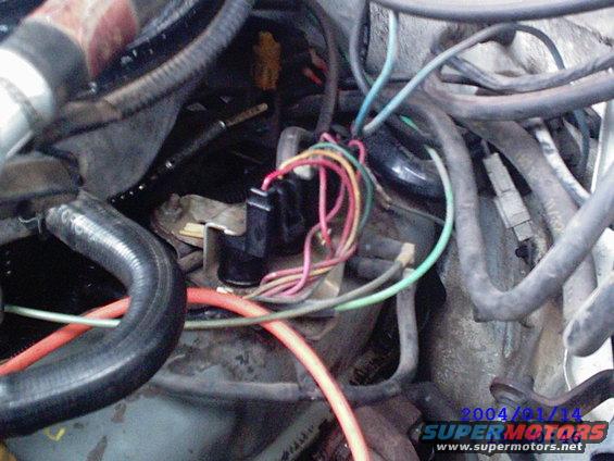 Engine bay with tangled wires and hoses. A black connector with multiple colored wires is visible