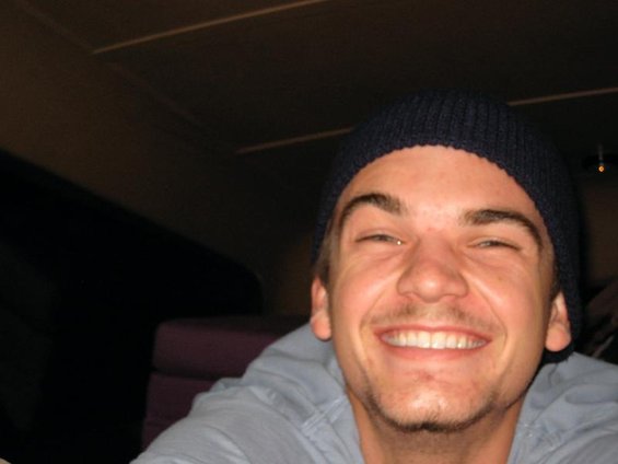 Man in a dark blue beanie and light blue shirt smiles widely, eyes squinting. Interior shot