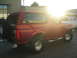 1995 Ford Bronco - A red 1995 Ford Bronco is seen from the rear quarter angle, with a spare tire mounted on the back. Another white vehicle is visible in the…