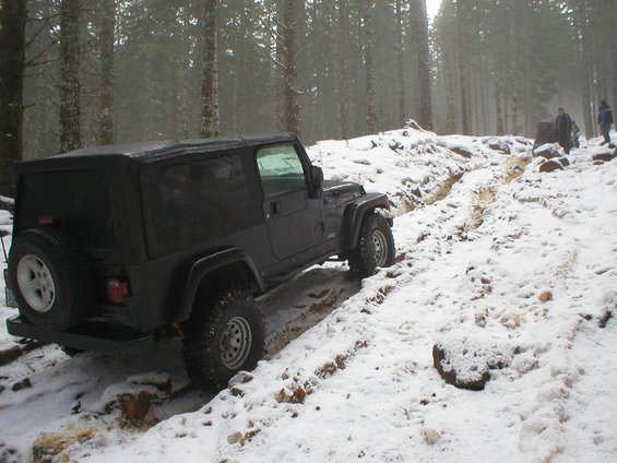 Black Jeep Wrangler driving on a snowy, wooded trail. The vehicle is angled on a rutted path with snow on the ground and trees in the background