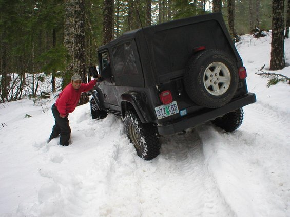 Black Jeep Wrangler with soft top driving in snow, person leaning on driver's door