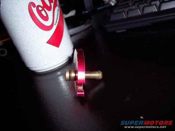 Red metal fuel rail adapter with brass fittings next to a Coca-Cola can