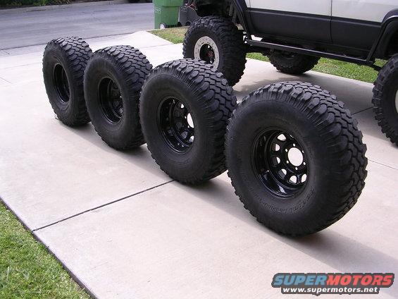 Four black off-road tires with aggressive tread patterns lined up on a concrete driveway, with a portion of a white and black off-road vehicle visible in the background