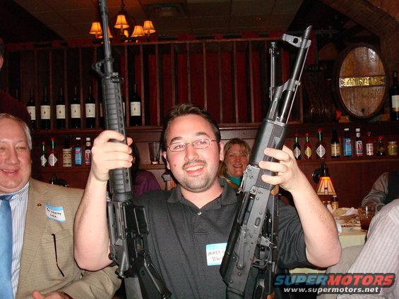 guy in black shirt holding AK-47, guy in tan jacket holding AR-15, people in background
