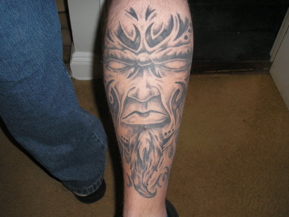 Tattoo of a tribal face with a beard on a leg, next to blue jeans