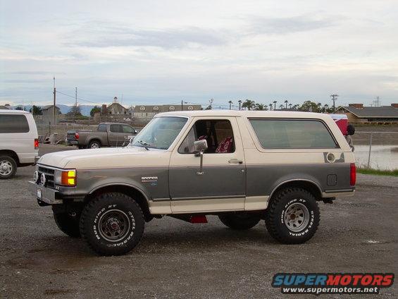 Two-tone Ford Bronco parked on gravel, side view. It has off-road tires and a lifted suspension