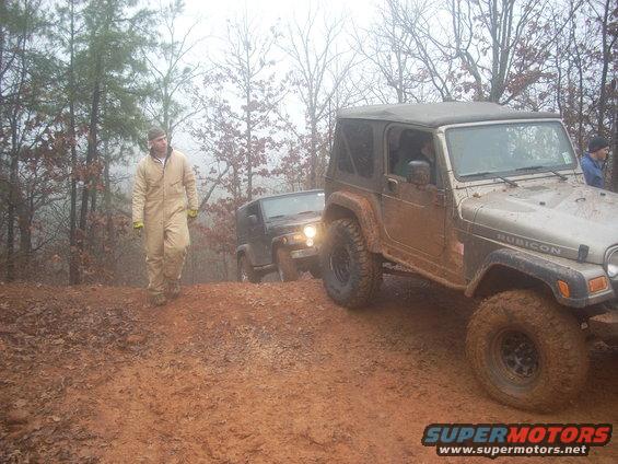 Two Jeeps, one tan and one black, driving off-road on a muddy incline with trees in the background