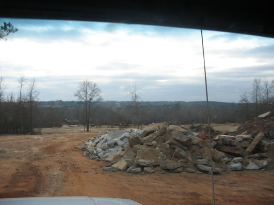 View from inside a vehicle looking out at a construction site with piles of rocks and debris, bare trees, and a cloudy sky