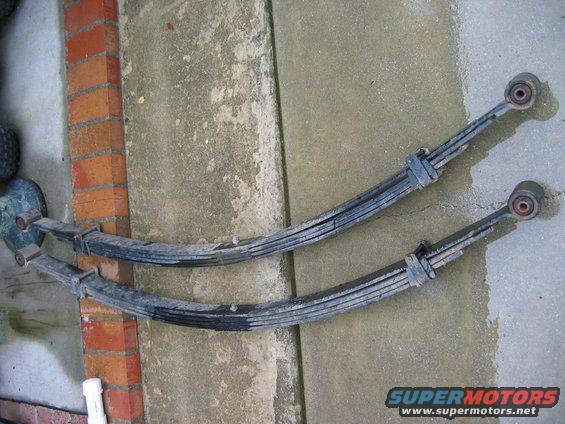 Two leaf springs are laid out on a concrete surface next to a brick wall. One spring is slightly above and behind the other