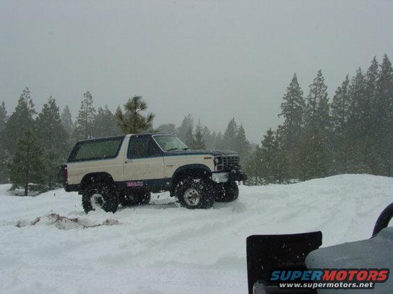 1980s Ford Bronco with off-road tires driving in snow, trees in background