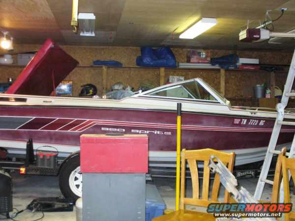 Boat with maroon stripe and 'sea sprite' text, parked in a garage with a ladder and chairs