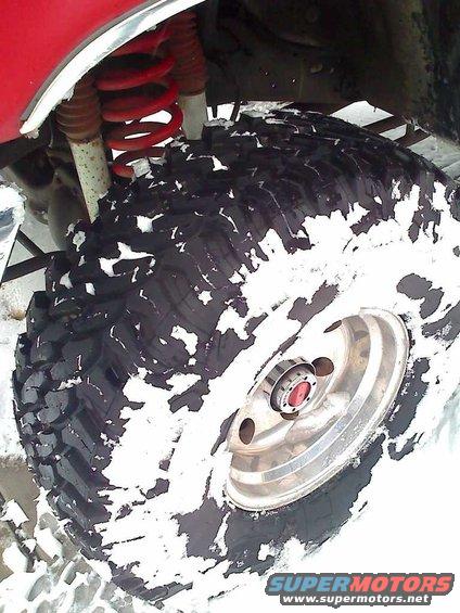 Close-up of a large off-road tire with snow on it, showing red suspension and part of a red vehicle body