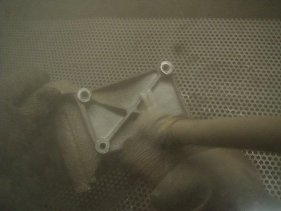Metal bracket with four bolt holes and a pipe attached, seen in a dusty environment