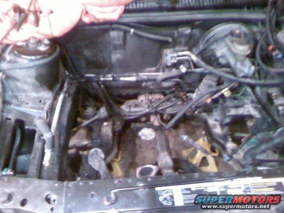 Engine bay with exposed engine components, hoses, and wiring. A yellow jack stand is visible