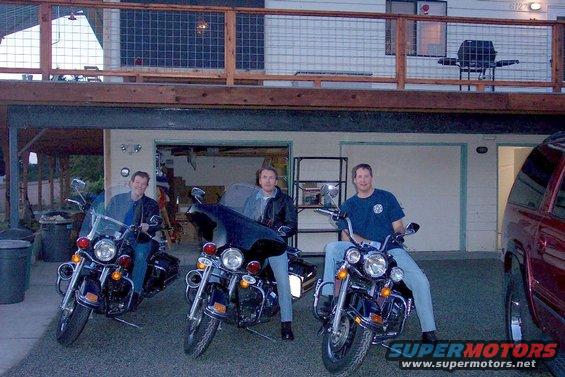 Three men sit on motorcycles in front of a garage and house