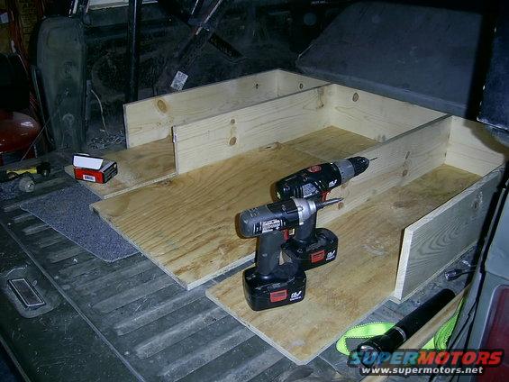 Wooden storage box being built inside a vehicle's cargo area, with two cordless drills on the plywood