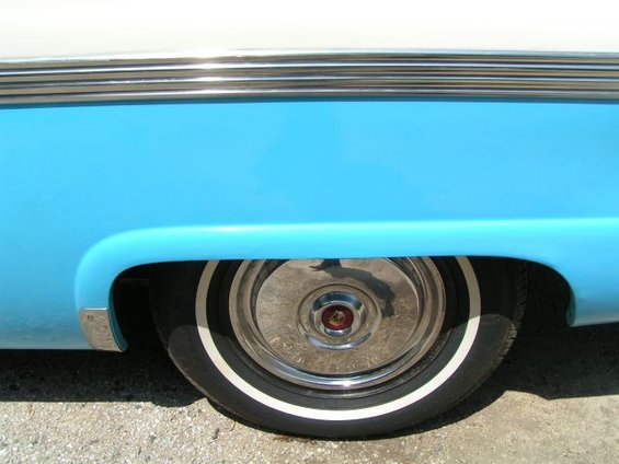 Close-up of a blue and white car's rear wheel well, showing a whitewall tire and chrome hubcap