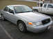 2004 Ford Crown Victoria - Silver sedan with a black grille and chrome wheel covers is parked in front of a building, with a pickup truck behind it.