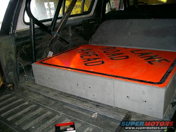 Interior view of a vehicle cargo area with a storage box topped by a 'Road Ahead Lane' traffic sign