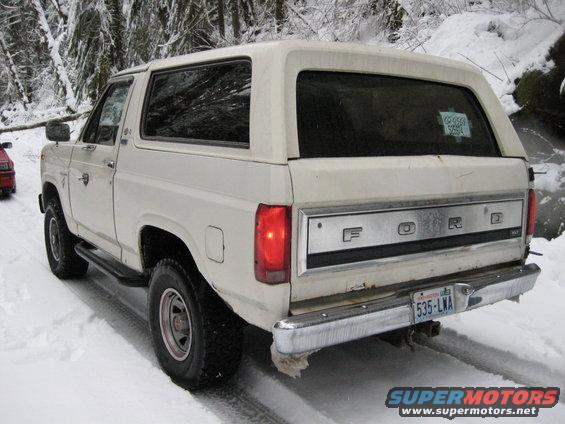 White 1980 Ford Bronco in snow, rear view, with a red car visible to the left