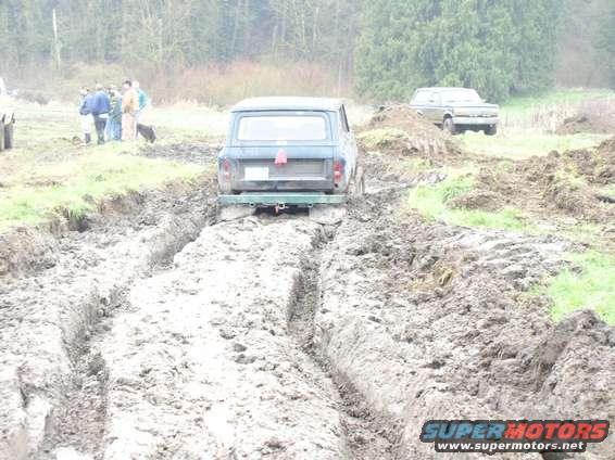Rear view of a blue Jeep CJ-5 stuck in mud, with another vehicle in the background