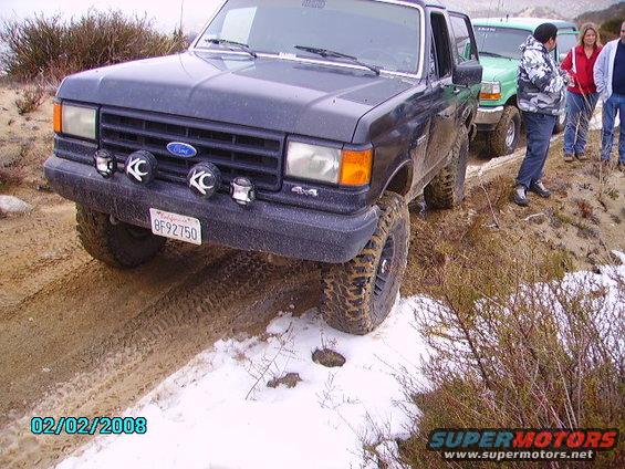 Dark gray Ford Bronco with KC lights and aftermarket wheels on a muddy trail with snow patches