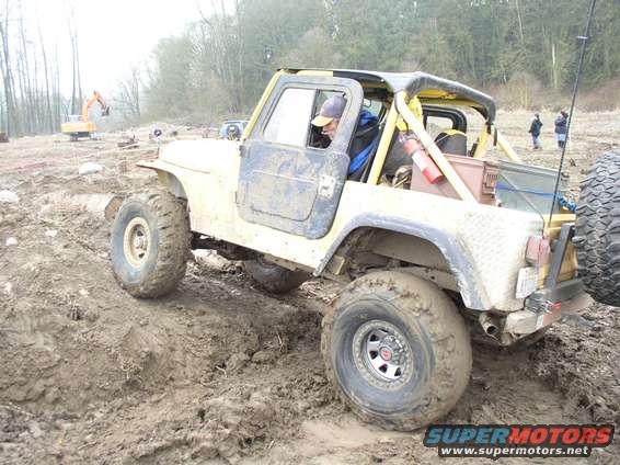 Yellow off-road vehicle with a roll cage driving through mud. Driver wearing a blue shirt and a baseball cap