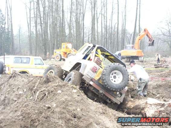 1978 Ford Bronco tilted in mud with large tires, yellow van and construction equipment in background