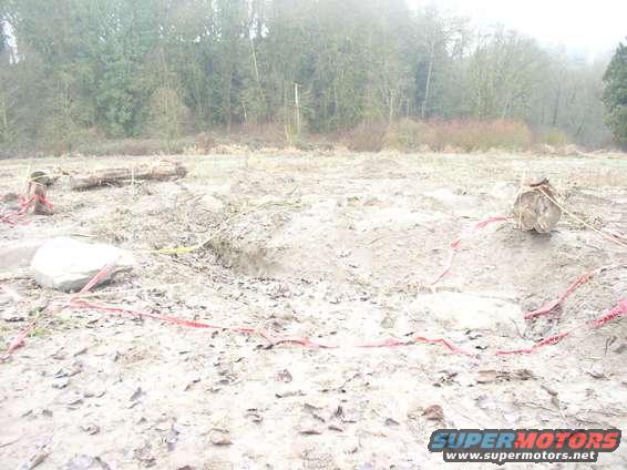 Construction site with logs, rocks, and red caution tape on muddy ground. Trees in background