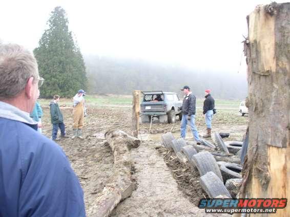 Off-road vehicle in a muddy area with people around. A second white vehicle is visible in the background